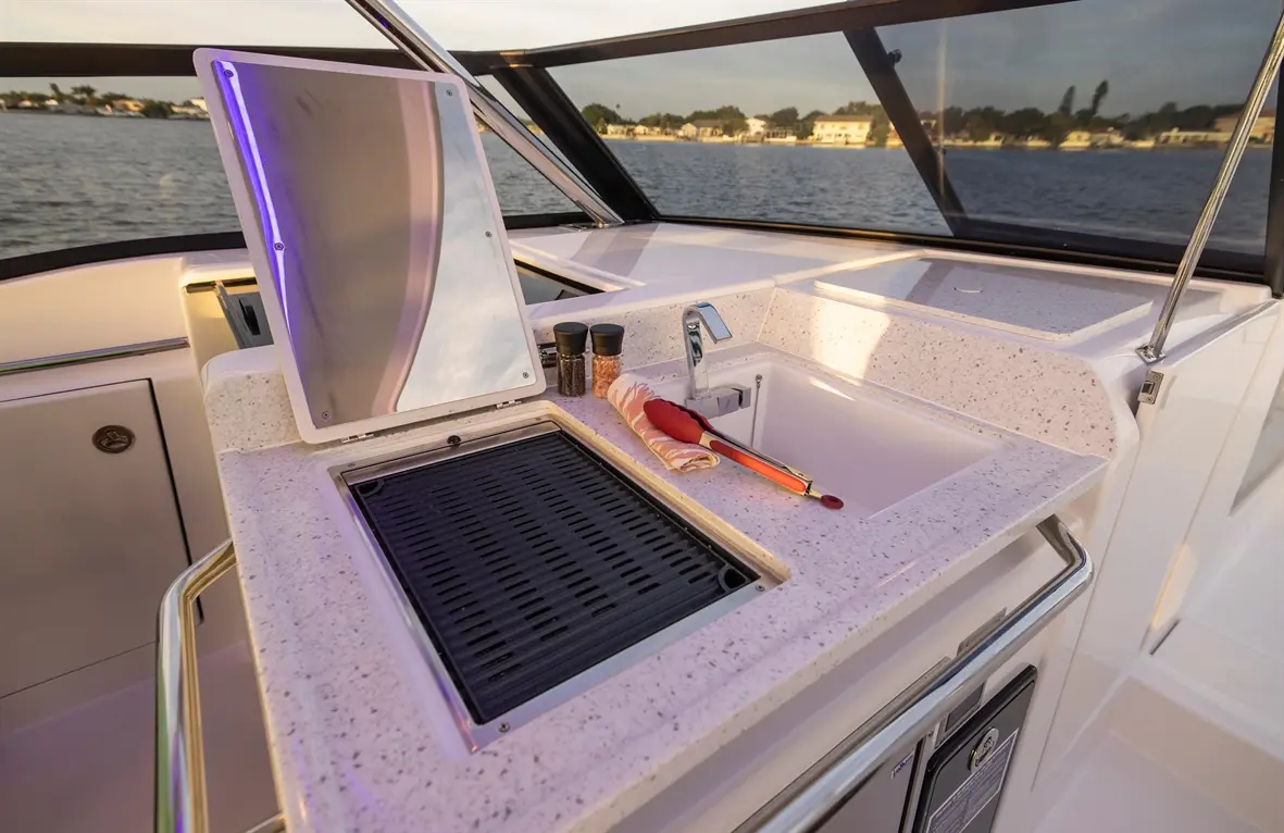 Modern boat kitchen with a grill, sink, spices, tongs, and a mirror, all set against a scenic waterside backdrop.